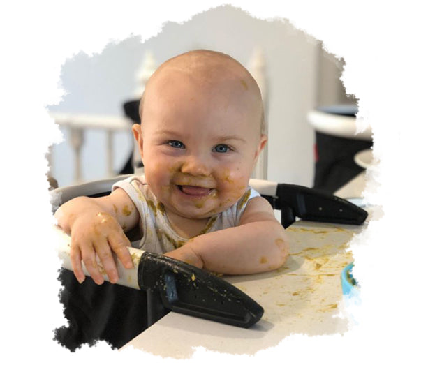 baby with messy hands and face smiles for the camera while feeding at the table in their lobster portable highchair - philandteds